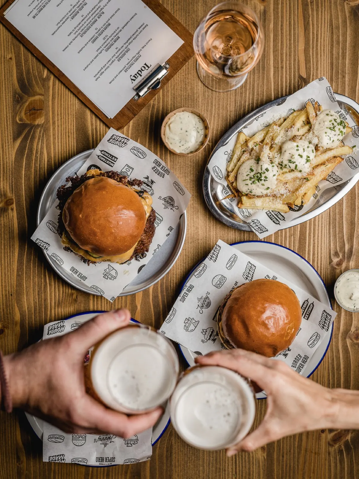 Table with two burgers, fries with sauce and beers clinking